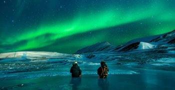 Two people sitting on frozen lake enjoying aurora borealis in Alaska